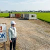 「資材置場OK」と書かれた貸地の看板の横で、ヘルメット姿の行政書士がスクラップヤード（特定再生資源屋外保管業）の新規開業に向けて現地の法令適合性を調査している様子。隣接する青々とした農地（田んぼ）やコンテナの存在から、農地法違反や市街化調整区域での無許可建築といった、土地契約前に確認すべきリスクを示唆している。